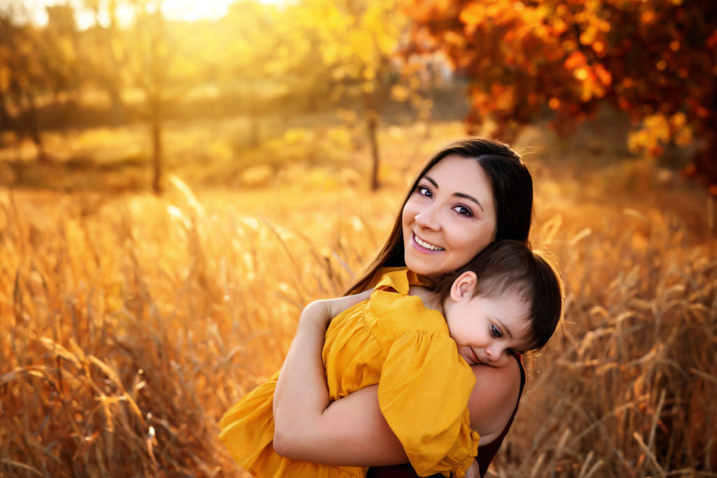 Woman carrying daughter with yellow dress, with the sun glowing the prairie and red leafed trees behind her