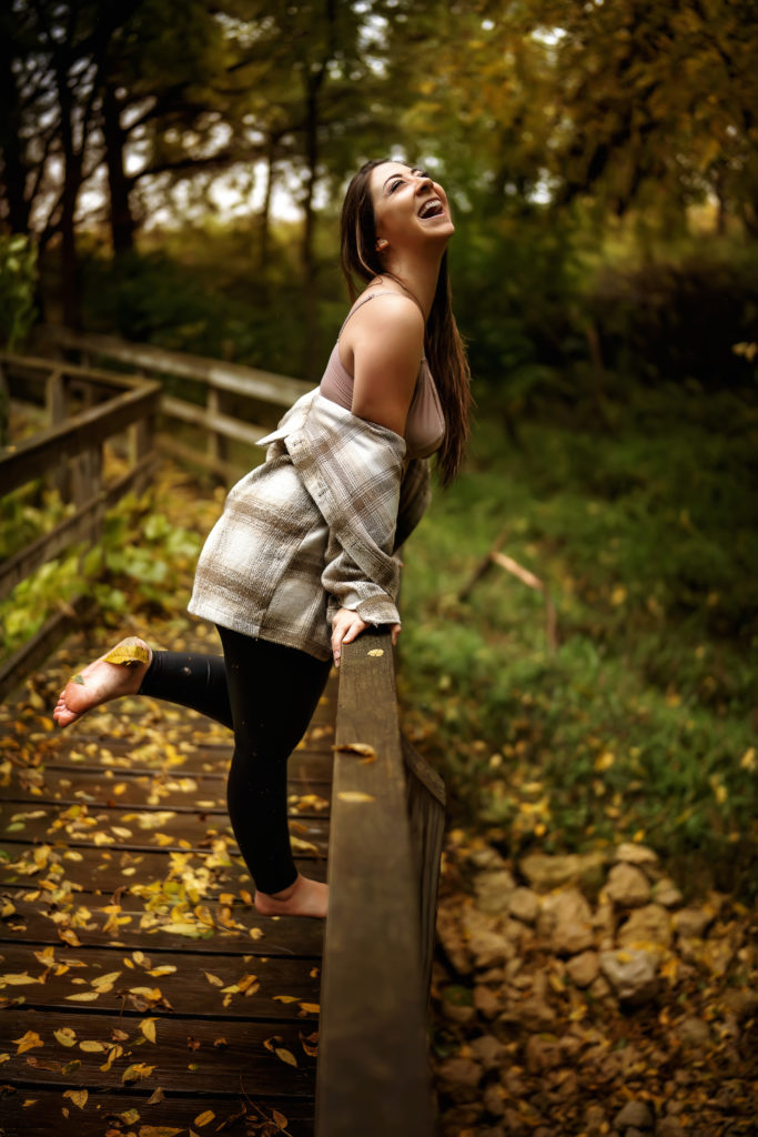 woman leaning on a leaf covered bridge in Illinois, jacket fall on her shoulders, smiling and laughing up into the rain