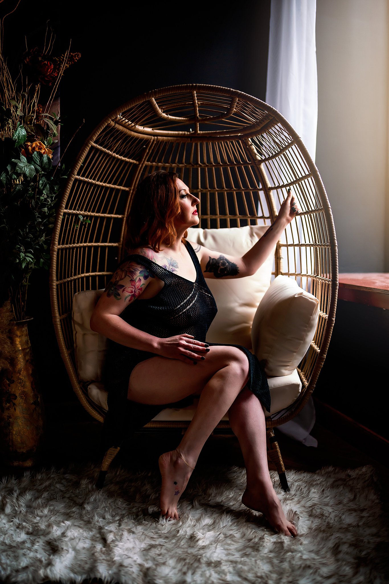 A woman with red hair sits in a wicker egg chair in black lingerie looking out the window before exploring Chicago Nightlife