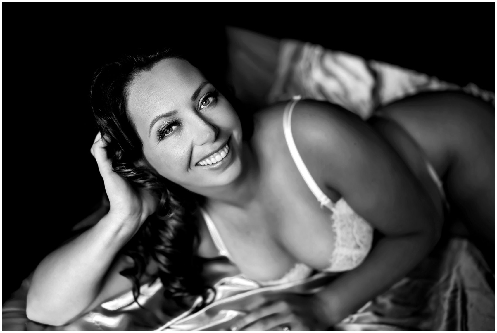 A smiling woman lays across a bed leaning on her elbow