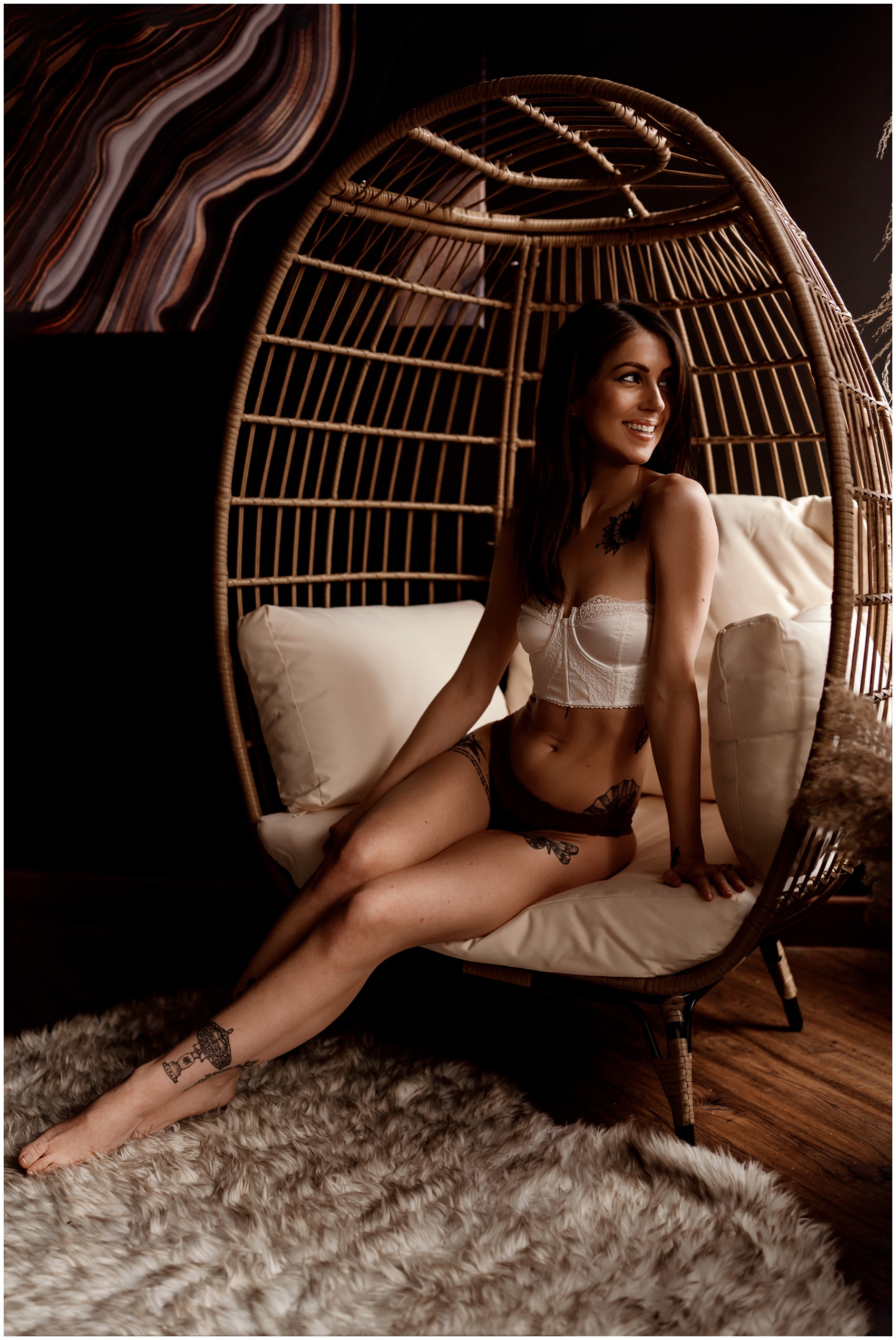 A smiling woman in a small white corset and black undies sits in a large egg wicker chair in a studio after getting lash extensions elgin