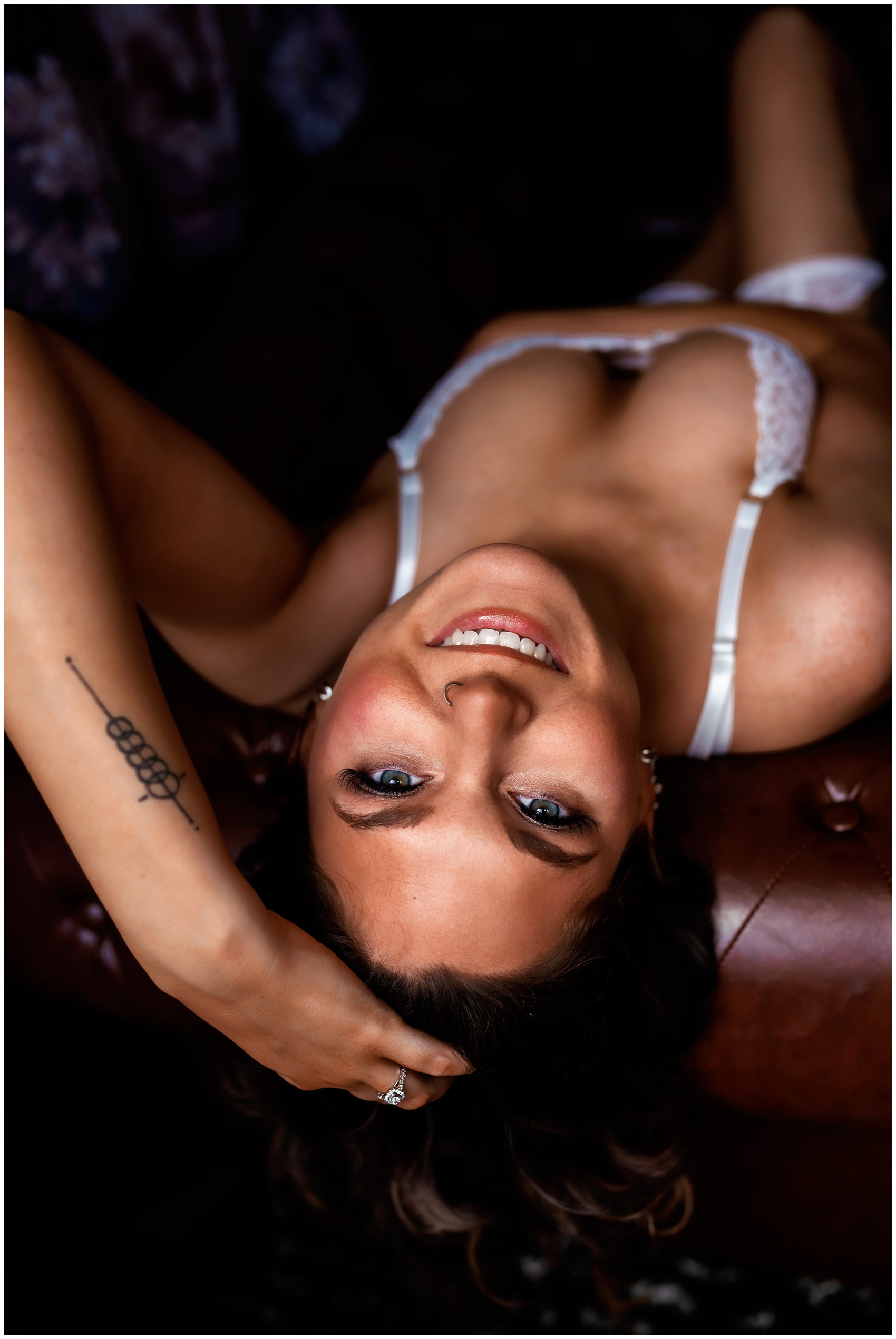 A woman smiles big while laying on a leather couch in white lace lingerie after some microblading elgin